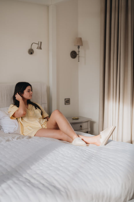 model wearing light coloured undyed slip on sheepskin slippers on a bed