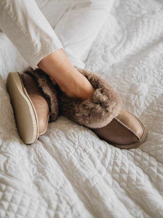 Sheepskin Slipper in Walnut and stone wool with a Napallan finish, worn by a woman on a white bed.