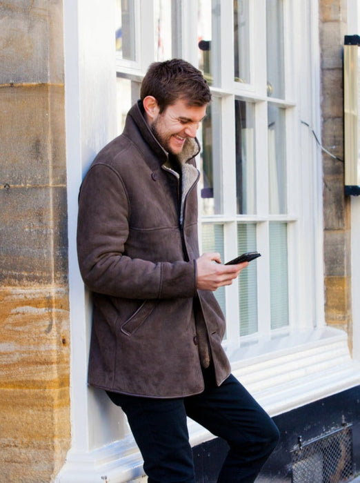 Men's Sheepskin Coat. Male model wears brown Sheepskin coat, with hand in pocket, looking down at phone.