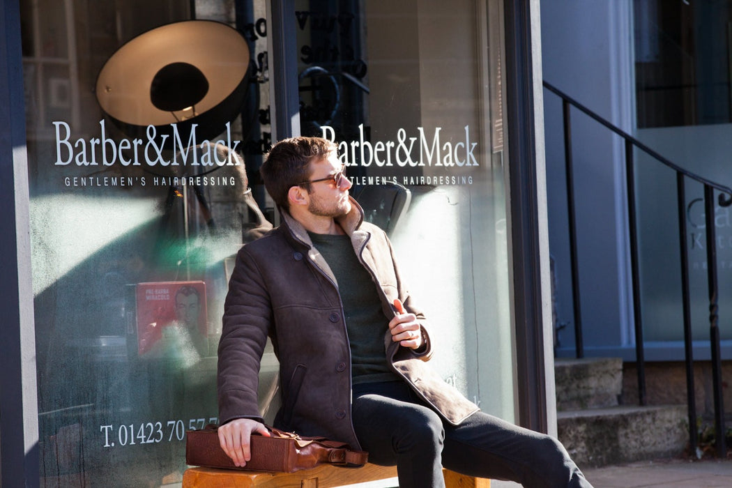 Male model holds open Men's Sheepskin brown Edward Coat, model looks into sunlight holding brown leather bag.