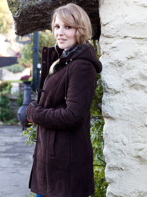 Women's Brown Sheepskin Duffle Coat, Millie. Model Wears Duffle coat with hood, stood outside thatched cottage.