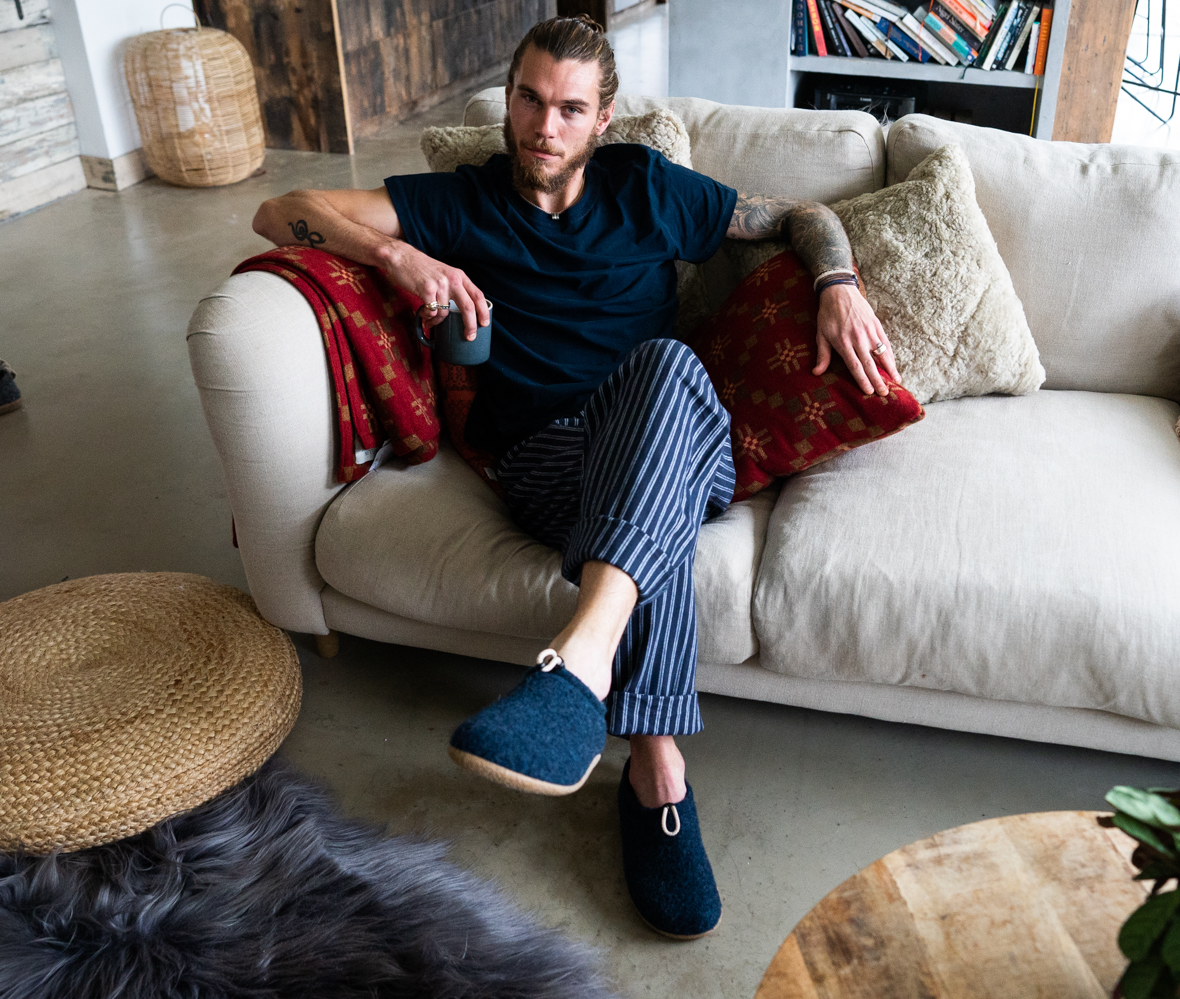 Male model sat on sofa wearing Mattias Slippers in Navy. Featured in the room is St Davids Cross Throw and Cushion in Chestnut. 