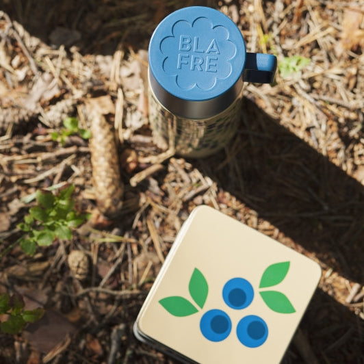 Blueberry Tin Fruit Pattern Box and Blueberry Fruit Print Vacuum Flask. Photographed on forest floor with pine cones and shrubbery.