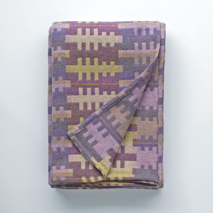 Forest woven wool blanket by Melin Tregwynt. A purple striped blanket with green and cream tree like shapes in repeating pattern. Images shows folded blanket with corner folded over to show reverse.