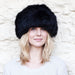 A blonde female model is smiling, she wears a large black Sheepskin hat. The hat is rounded and covers her full head just above her eyebrows.