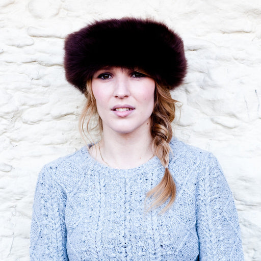 A blonde female model with her hair in a side plait wears a light blue cable knit jumper.. She wears a rich Brown full Sheepskin headband. The headband covers her forehead and ears.