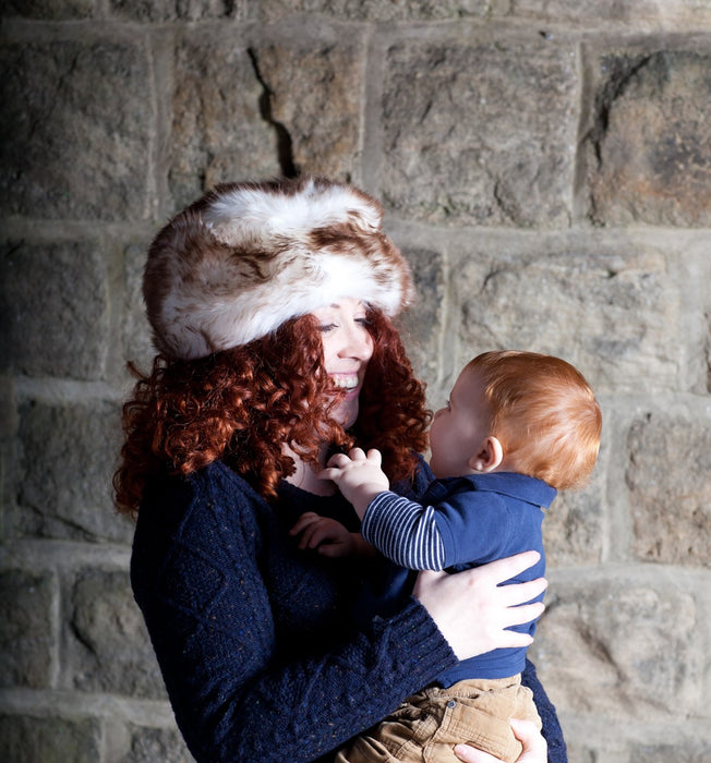 An auburn female model is smiling, holding a laughng child. She wears a dark Navy cable knit jumper. On her head she wears a Natural White and Brown full Sheepskin Hat. 