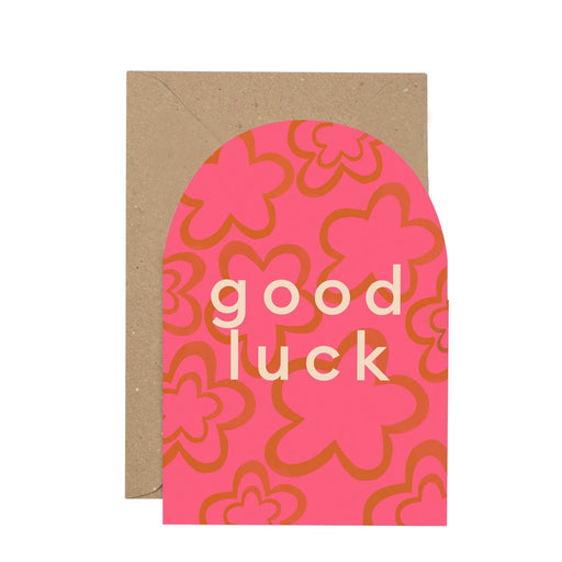 Good Luck greetings card. A hot pink curved top verticle rectangular greetings card. Features mustard yellow flower shapes with white writing in the centre reading 'good luck' in large letters. Comes with an envelope.
